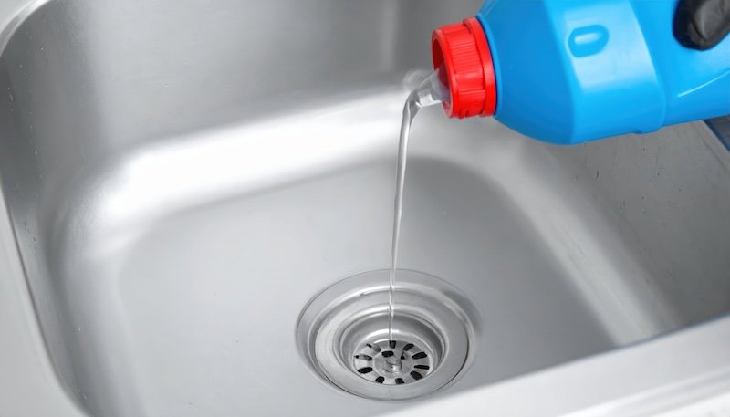 A close-up view of a person pouring a chemical drain cleaner into a kitchen sink to efficiently unclog drains, showcasing home maintenance techniques.
