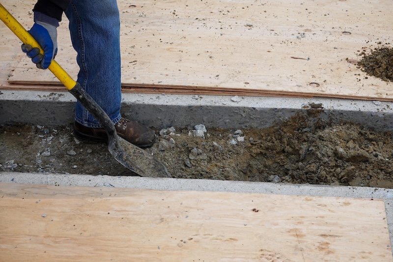 Leg of laborer with shovel digging a ditch through a channel between concrete slabs