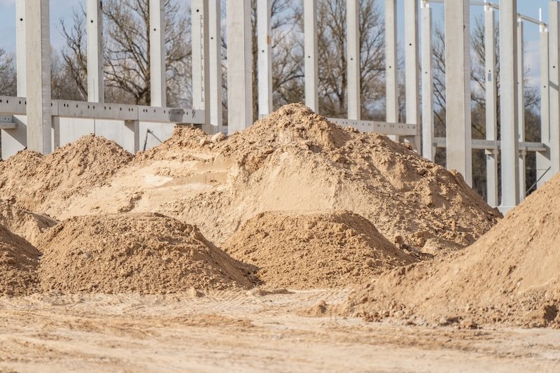 Large piles of sand and aggregate staged on warehouse construction site beside concrete column grid for earthworks, grading, and material supply use