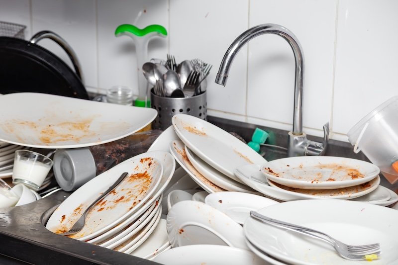 Large metal sink with dirty dishes in professional restaurant kitchen, pile of unclean white plates, crockery, appliances with leftover food, water tap, tiled wall
