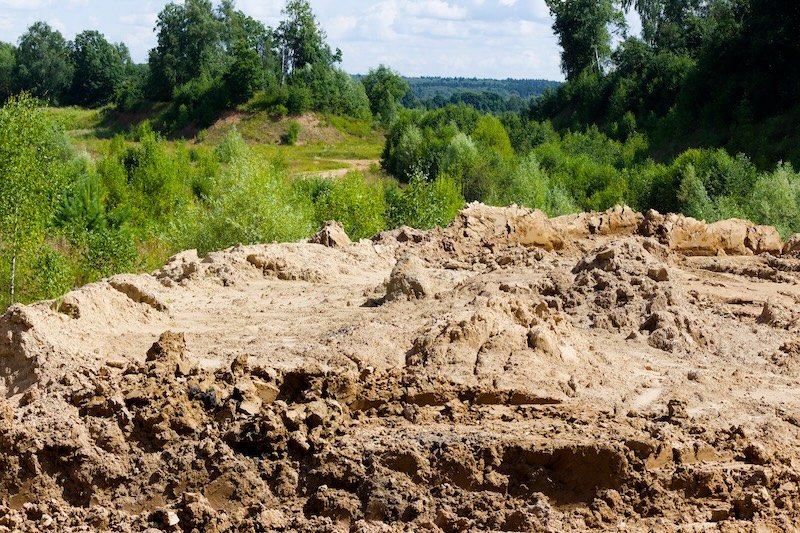Large pile of excavated soil and clay forming a rustic dirt mound at a construction site with green forest and trees growing in the scenic background under a summer sky