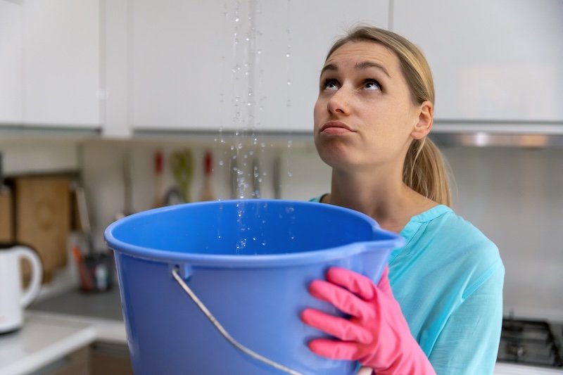 home flooded by upstairs or roof damage - woman holding bucket while water leaking from ceiling in kitchen hidden leaks