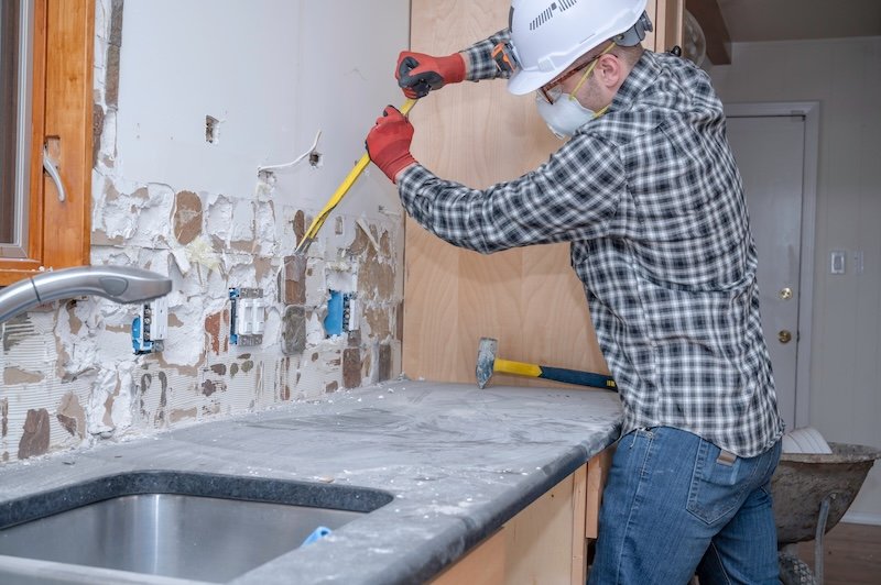 A renovation worker removing kitchen wall tile during a home improvement project.