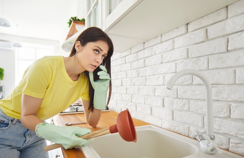 Worried young woman in rubber gloves holding cup plunger and calling plumber while standing in kitchen. Concerned female trying to fix clogged sink and solve home plumbing problem during housework.