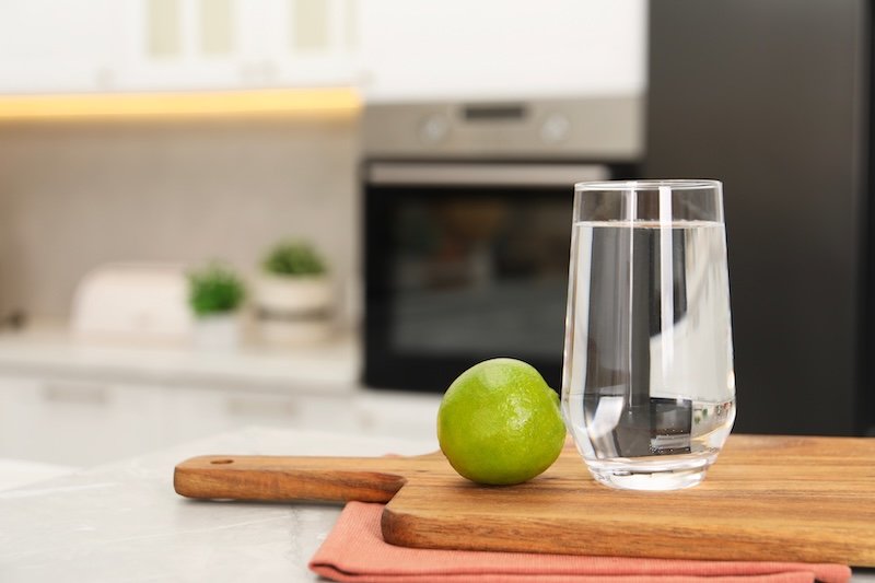 Filtered water in glass and lime on light marble table in kitchen, closeup. Space for text